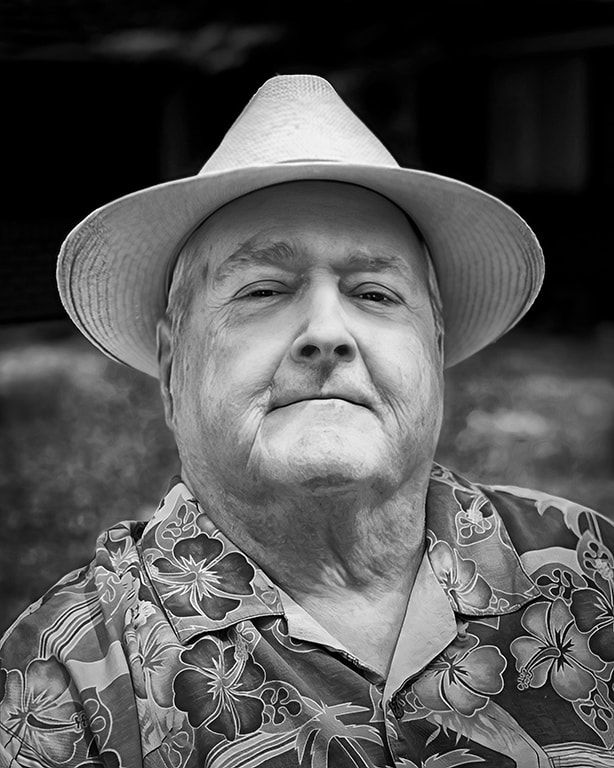 Portrait of a man in a light hat and floral shirt, looking at the camera with a neutral expression.
