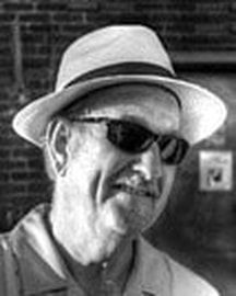 Black-and-white portrait of a man wearing a light fedora and dark sunglasses, smiling slightly.