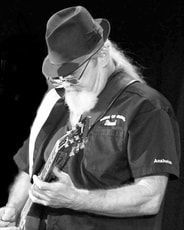 Black-and-white photo of a long-haired musician in a hat playing electric guitar onstage