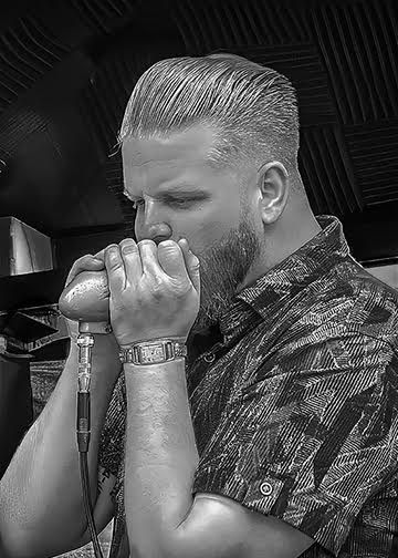 Man playing harmonica into a microphone, wearing a patterned shirt in a dark indoor setting