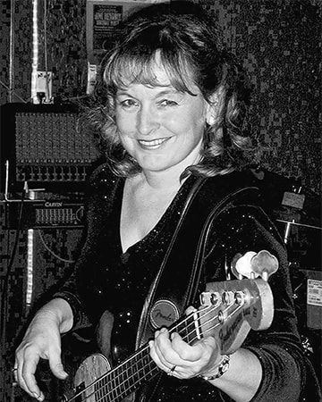 Smiling musician holding a bass guitar in a recording studio, with audio equipment behind her