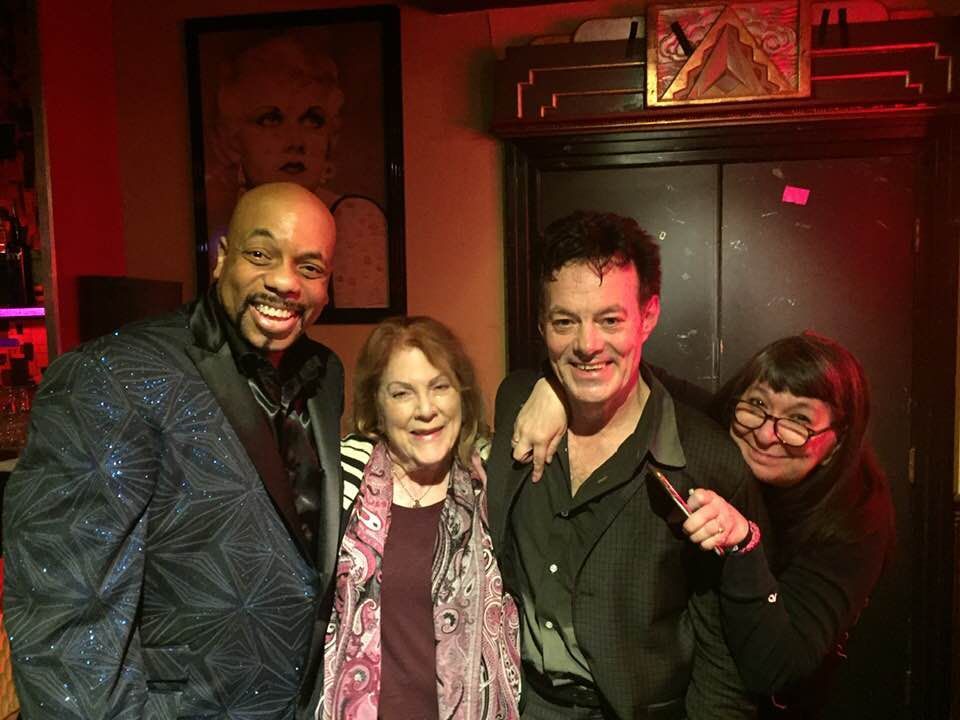 Four people smiling together in a dimly lit red-toned room, posing close with arms around each other.