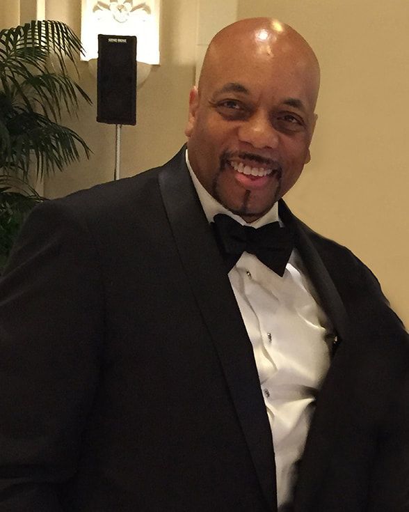 Smiling man in a black tuxedo and bow tie at an indoor event.