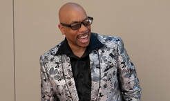 Man in glasses laughing, wearing a black shirt and patterned silver jacket against a beige wall.