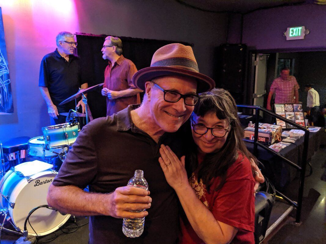 Two people smiling and hugging at a live music venue, one holding a water bottle.