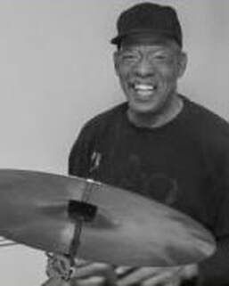 Smiling man in a cap playing drums, with a cymbal in the foreground.