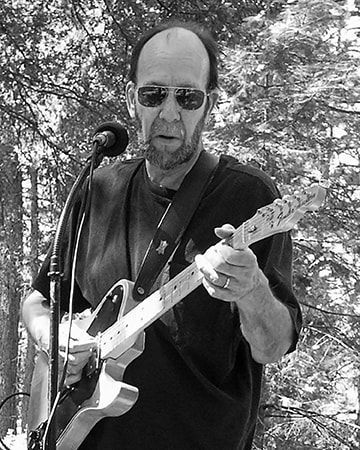 Man playing electric guitar outdoors, wearing sunglasses and a dark shirt.
