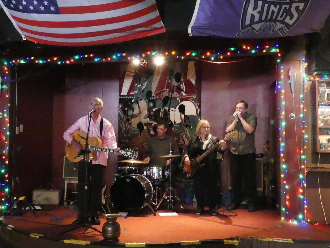 Four musicians performing on a small stage with string lights, drums, guitars, and microphones