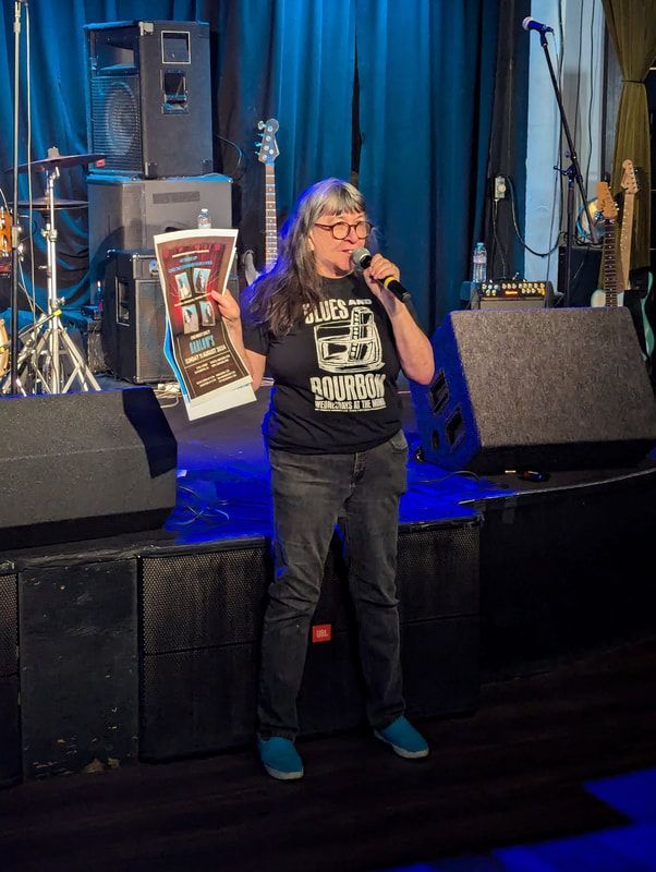 Person speaking on a stage, holding a framed sign, with blue curtains and concert equipment behind.