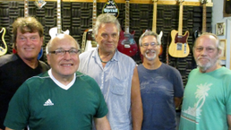 Five smiling people stand in a guitar shop with rows of guitars hanging behind them.