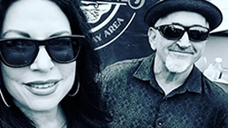 Two people wearing sunglasses pose in front of a circular sign.