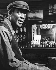 Black-and-white photo of a man in a cap smiling at a bar counter with bottles in the background
