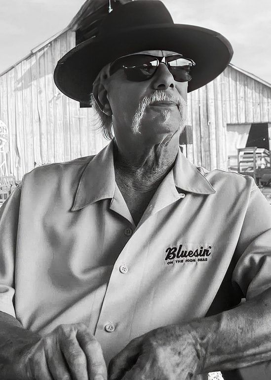 Man in a wide-brim hat and sunglasses, wearing a work shirt, leaning against a railing outdoors.