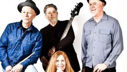 Four people posing against a white background, one holding a guitar, with a smiling woman in front.