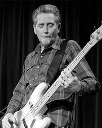 Man playing an electric bass guitar on stage, wearing a plaid shirt in black and white.