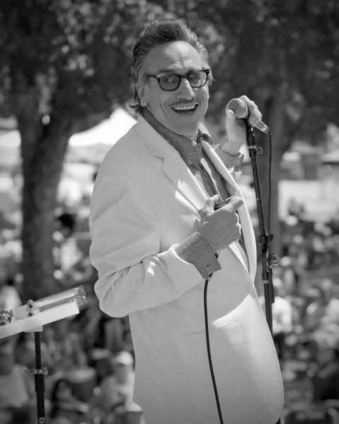 Smiling man in a light suit holding a microphone outdoors on a stage