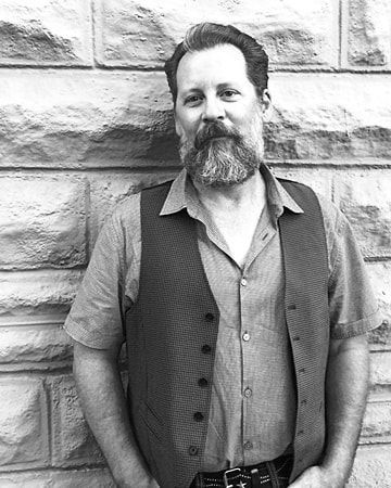 Man standing against a stone wall, wearing a vest and open-collared shirt, with hands in his pockets.