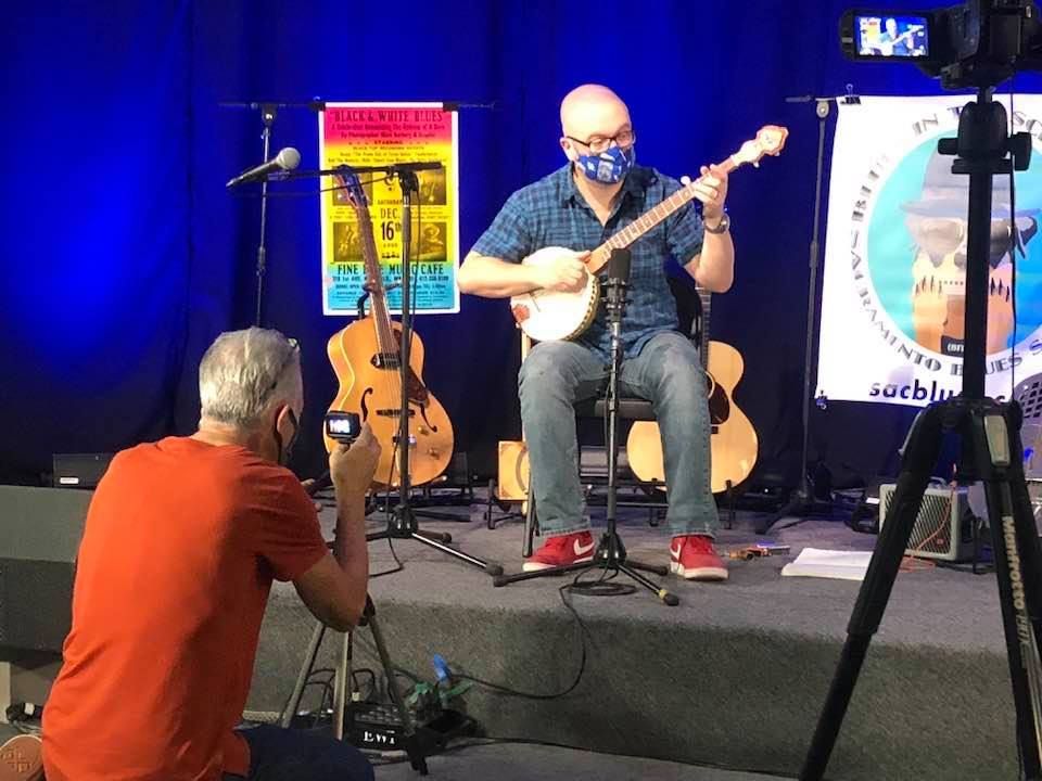 Musician seated on stage playing banjo, with guitars and blue lighting; photographer kneels in foreground.