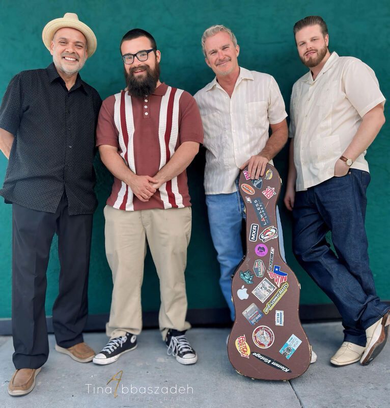Four men pose against a teal wall, one holding a sticker-covered guitar case.