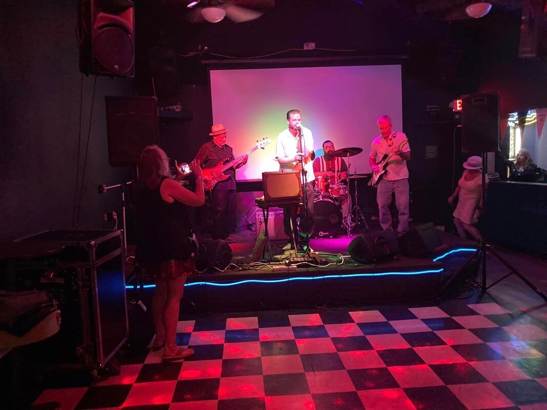 Live band performing on a small stage in a dark club with pink lighting and a checkered dance floor.
