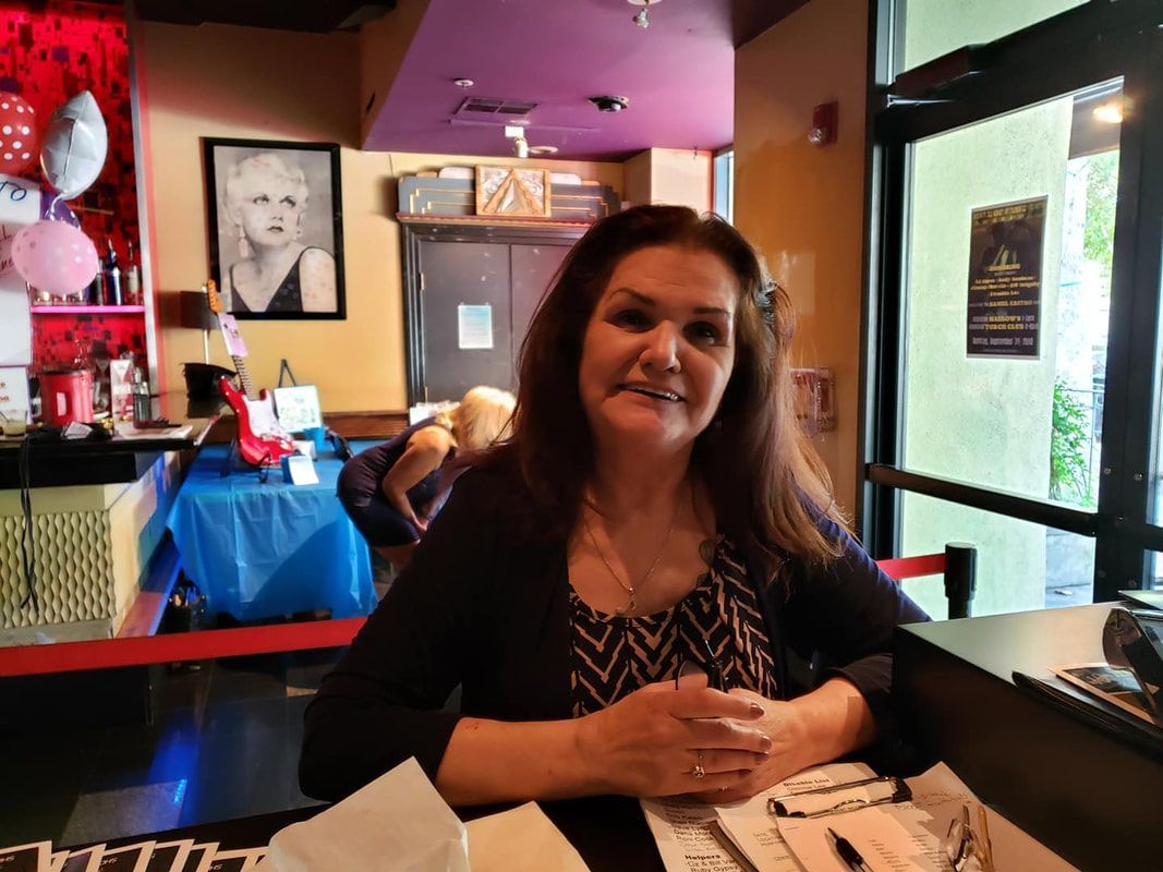 Smiling woman seated at a restaurant table with menus, near a doorway and colorful wall decor.
