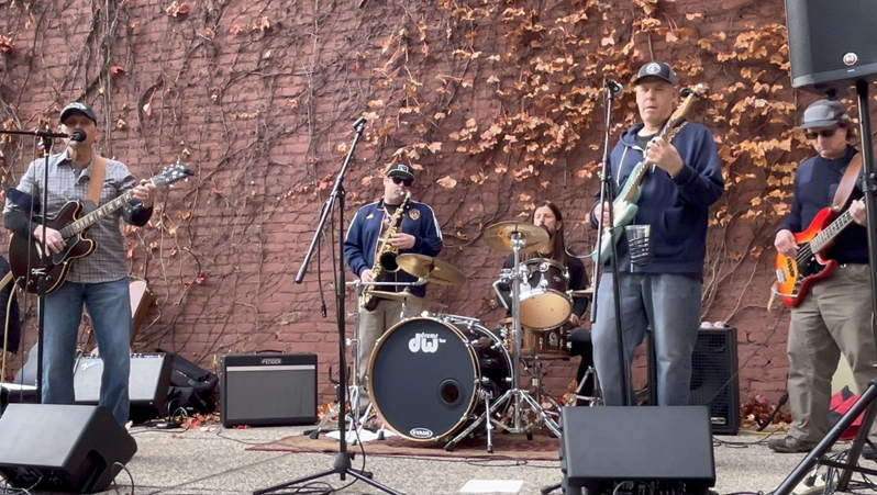 Band performing on an outdoor stage in front of a rocky wall, with guitars, drums, and microphones.