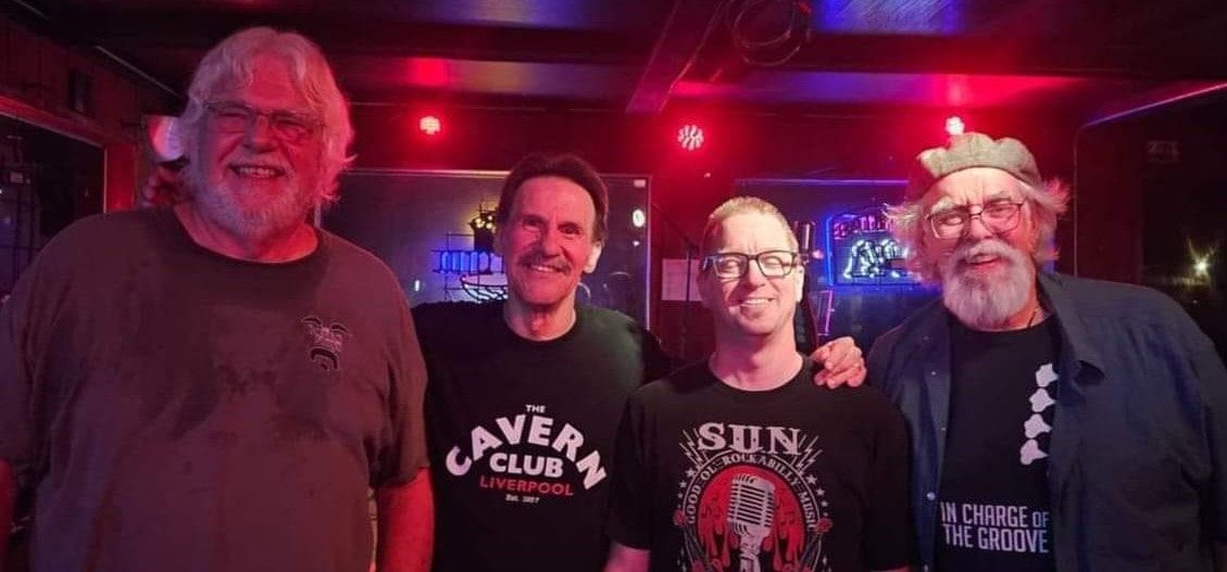 Four smiling people posing together in red stage lighting, wearing graphic T-shirts.