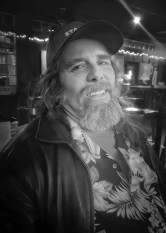 Black-and-white portrait of a smiling bearded man in a cap and leather jacket in a dimly lit bar.