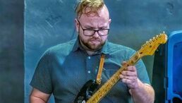 Man playing electric guitar onstage, wearing glasses and a blue shirt, focused on his instrument