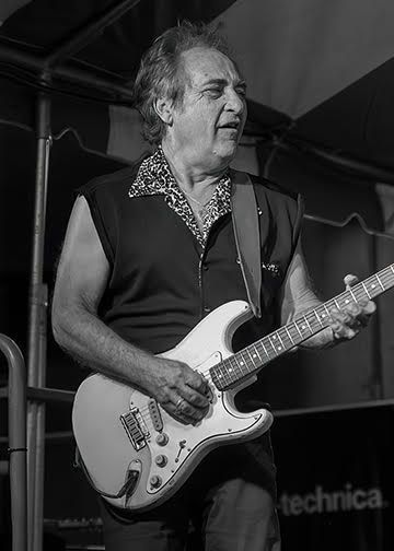 Musician playing an electric guitar on stage under a tent, wearing a dark vest and patterned shirt.