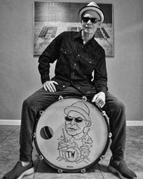 Man posing with a drum featuring a cartoon portrait, wearing a hat and sunglasses, in a room with framed art.