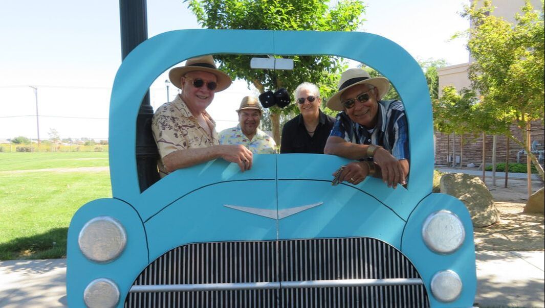 Three people posing in a bright blue vintage car cutout outdoors, smiling in sunglasses and hats