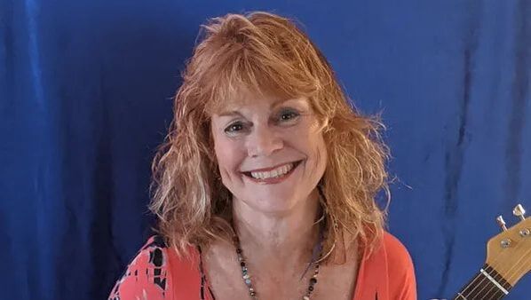 Smiling woman with curly red hair in a coral top, standing before a blue backdrop with a guitar neck visible