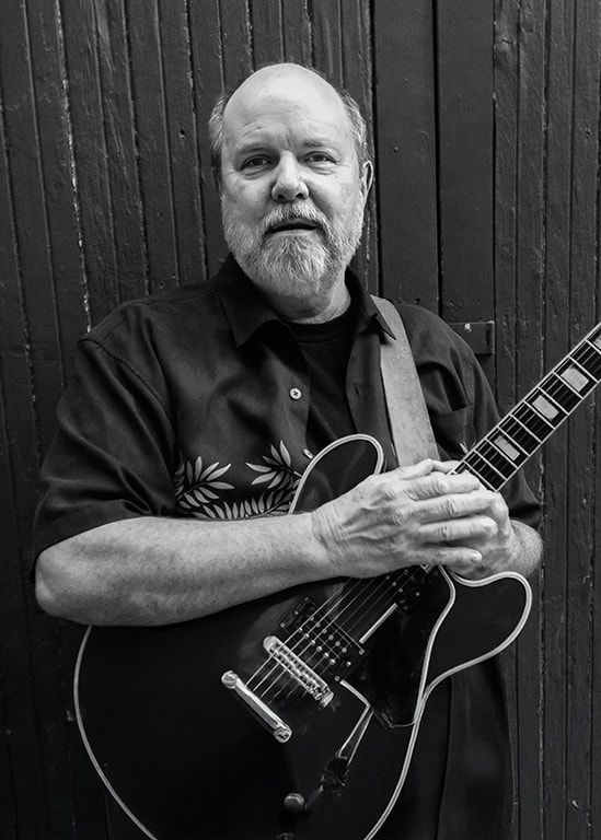 Man holding a guitar in front of a dark wooden wall, wearing a dark shirt and looking at the camera