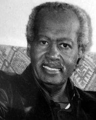 Black-and-white portrait of a smiling man seated against a patterned background