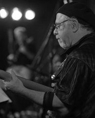 Black-and-white side profile of a person in a cap and glasses reading from papers onstage