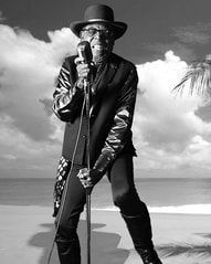 Singer in a black hat and leather jacket performing with a microphone on a beach backdrop.