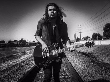 Black-and-white photo of a guitarist standing on railroad tracks holding an electric guitar outdoors