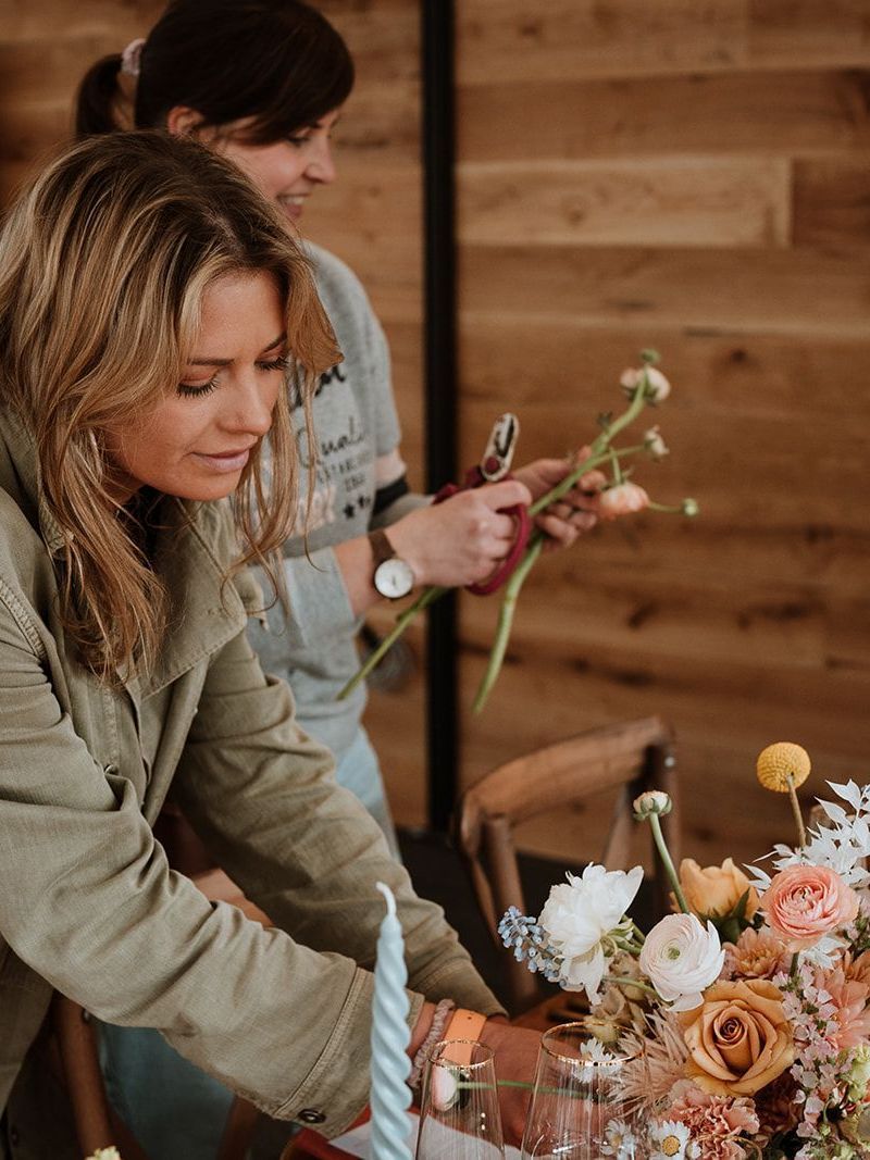 Two people arranging a floral centerpiece with candles on a wooden table in a rustic room