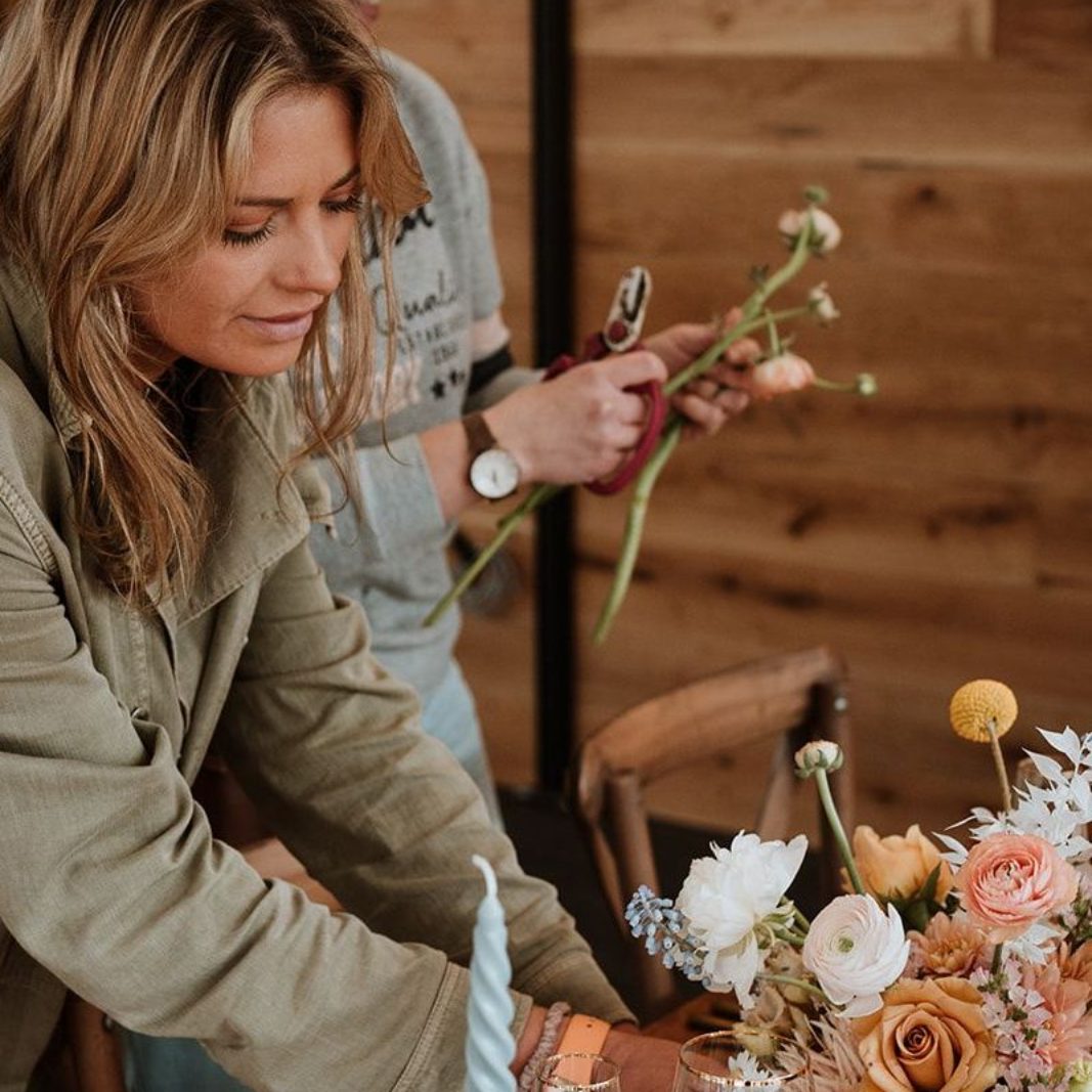 Person arranging a bouquet of white and peach flowers at a wooden table indoors