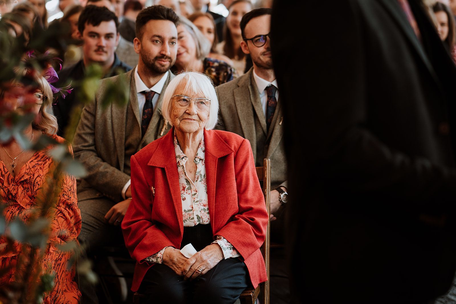 Elderly woman in a red jacket seated at an indoor event, with people standing behind her.