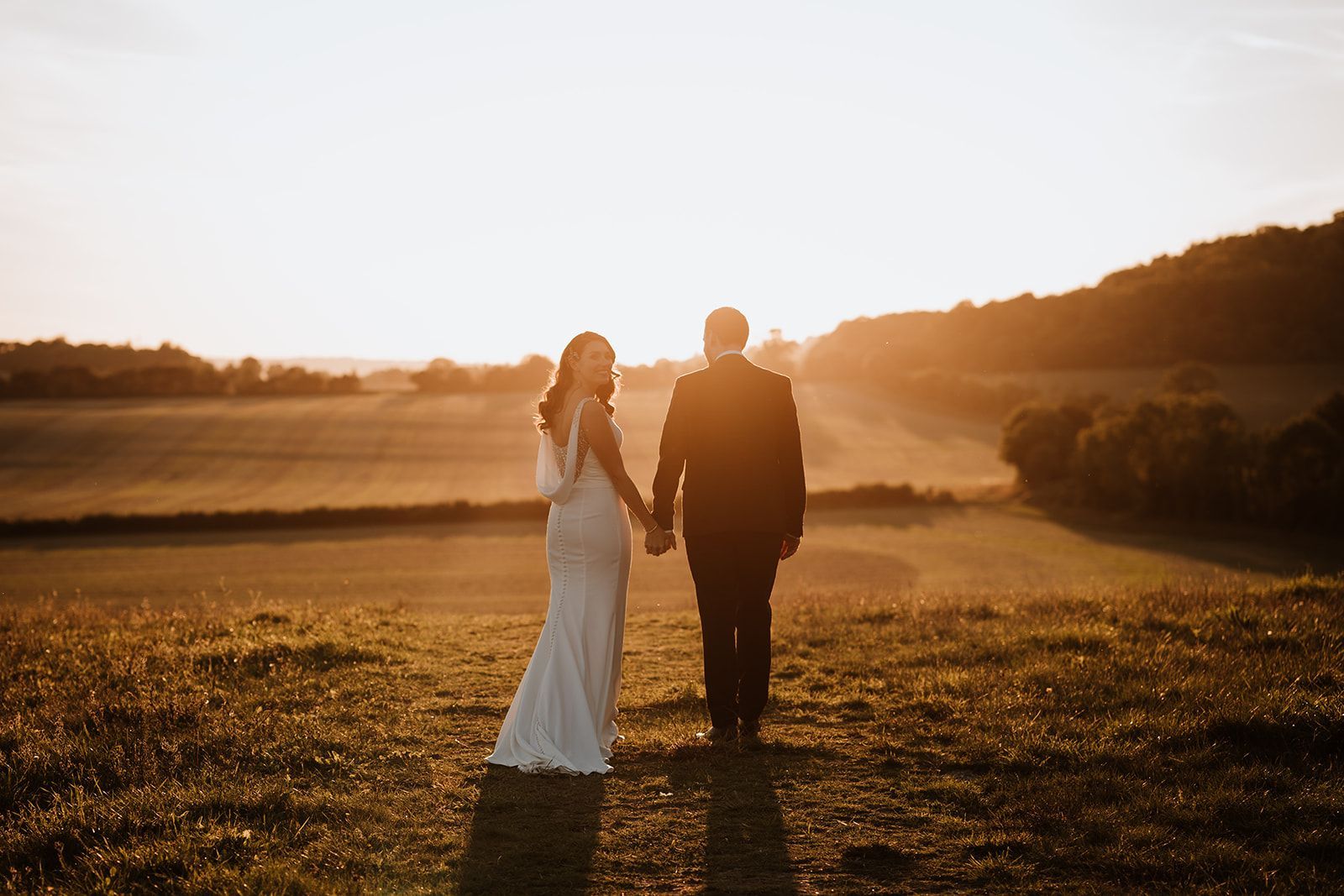 A couple holding hands walks through a sunlit field at sunset, with long shadows and warm golden light.
