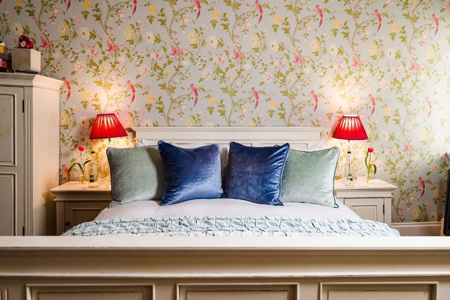 Cozy bedroom with a white bed, blue pillows, red lamps, and floral wallpaper