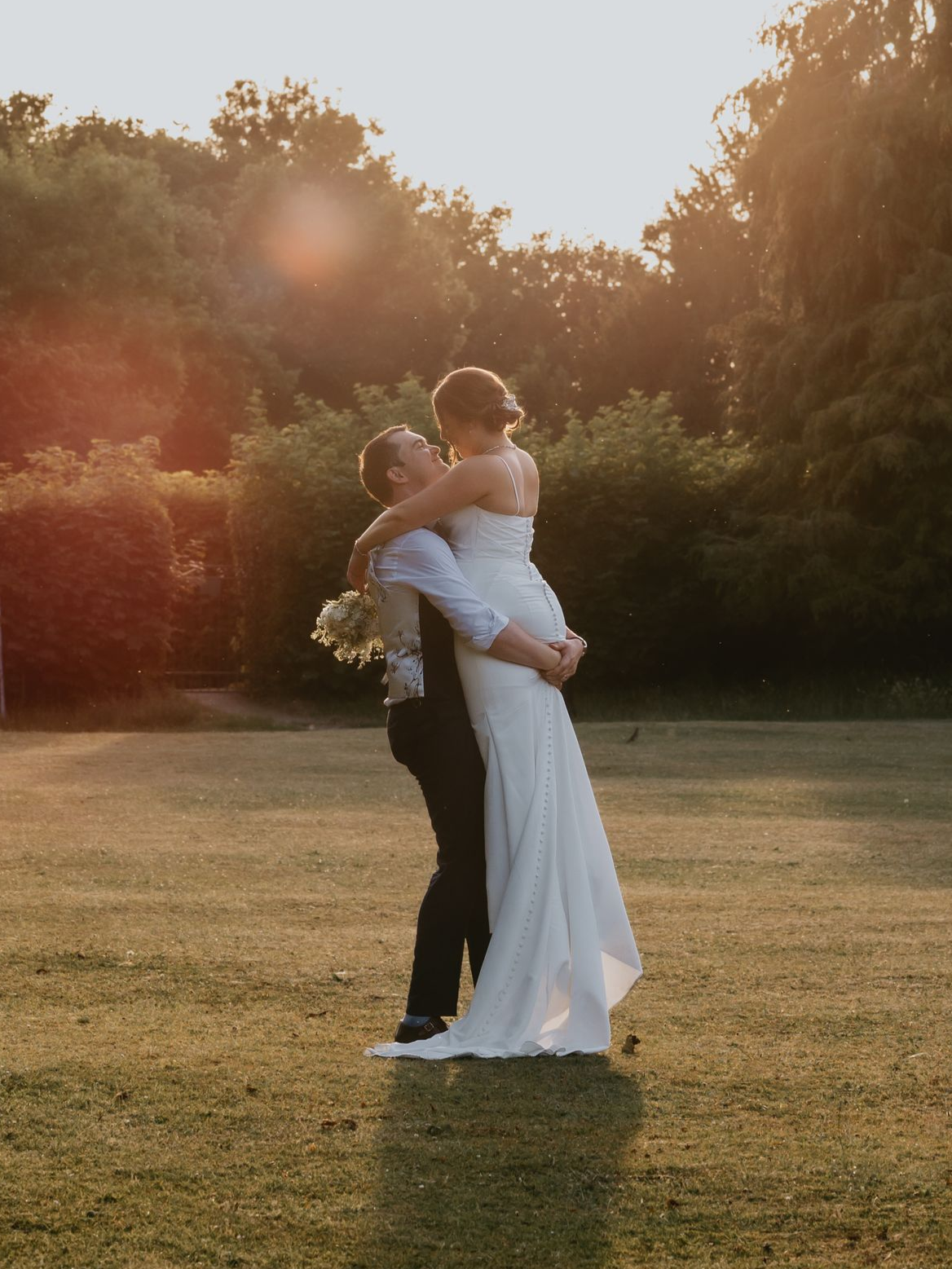 Two people embracing in a sunlit field, one in a white dress holding flowers, at sunset