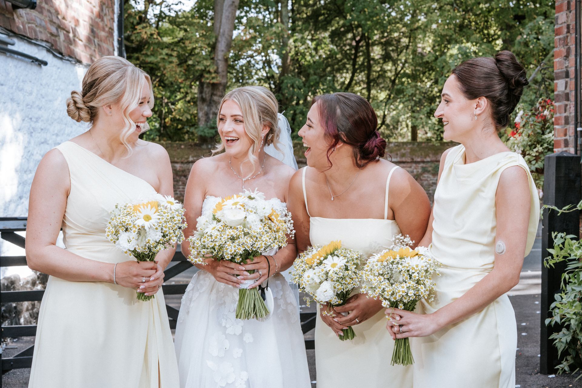 Four women in formal dresses holding bouquets and smiling outdoors at a wedding