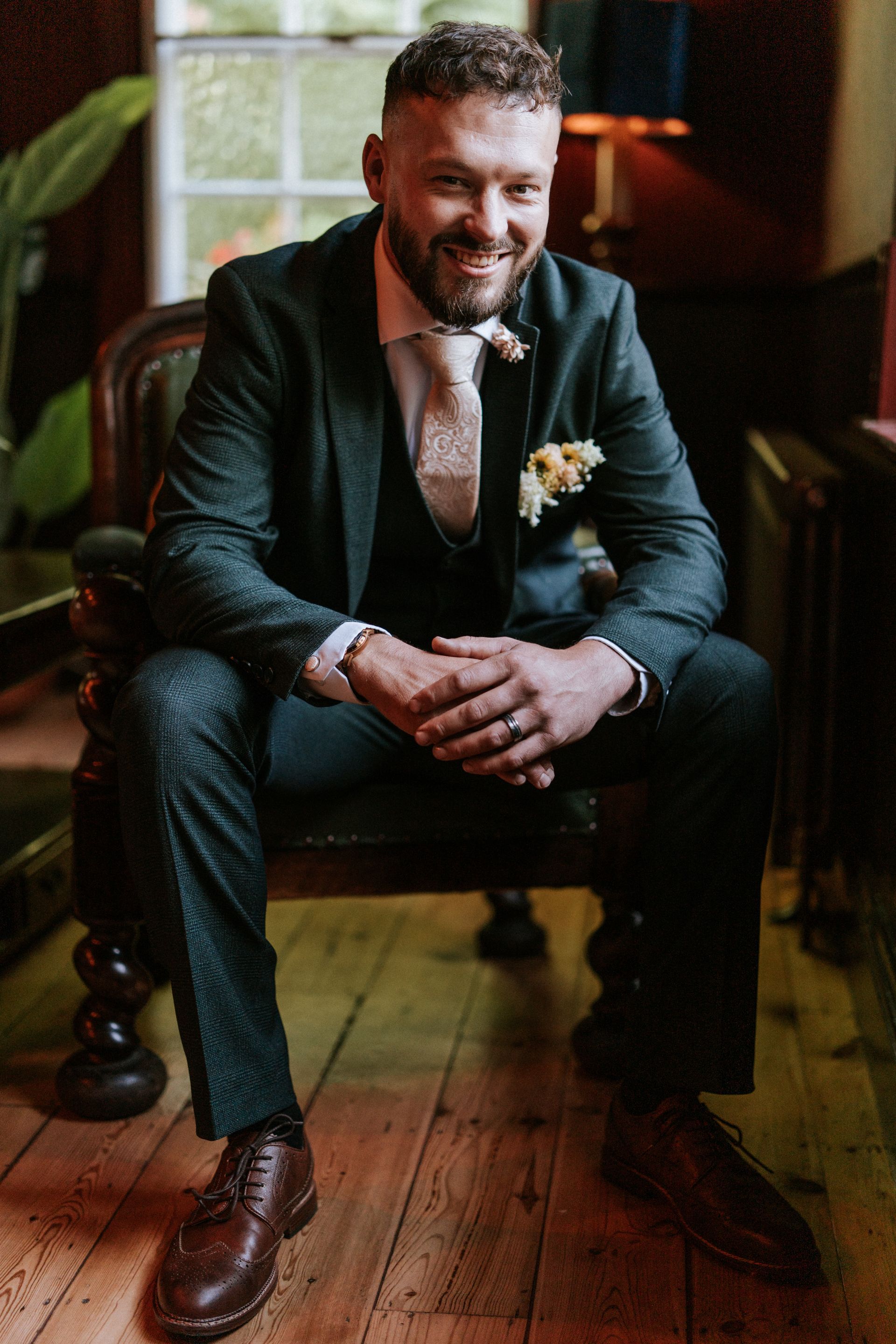 Smiling man in a dark suit sits on a chair in a warmly lit room, hands clasped, wearing brown shoes.