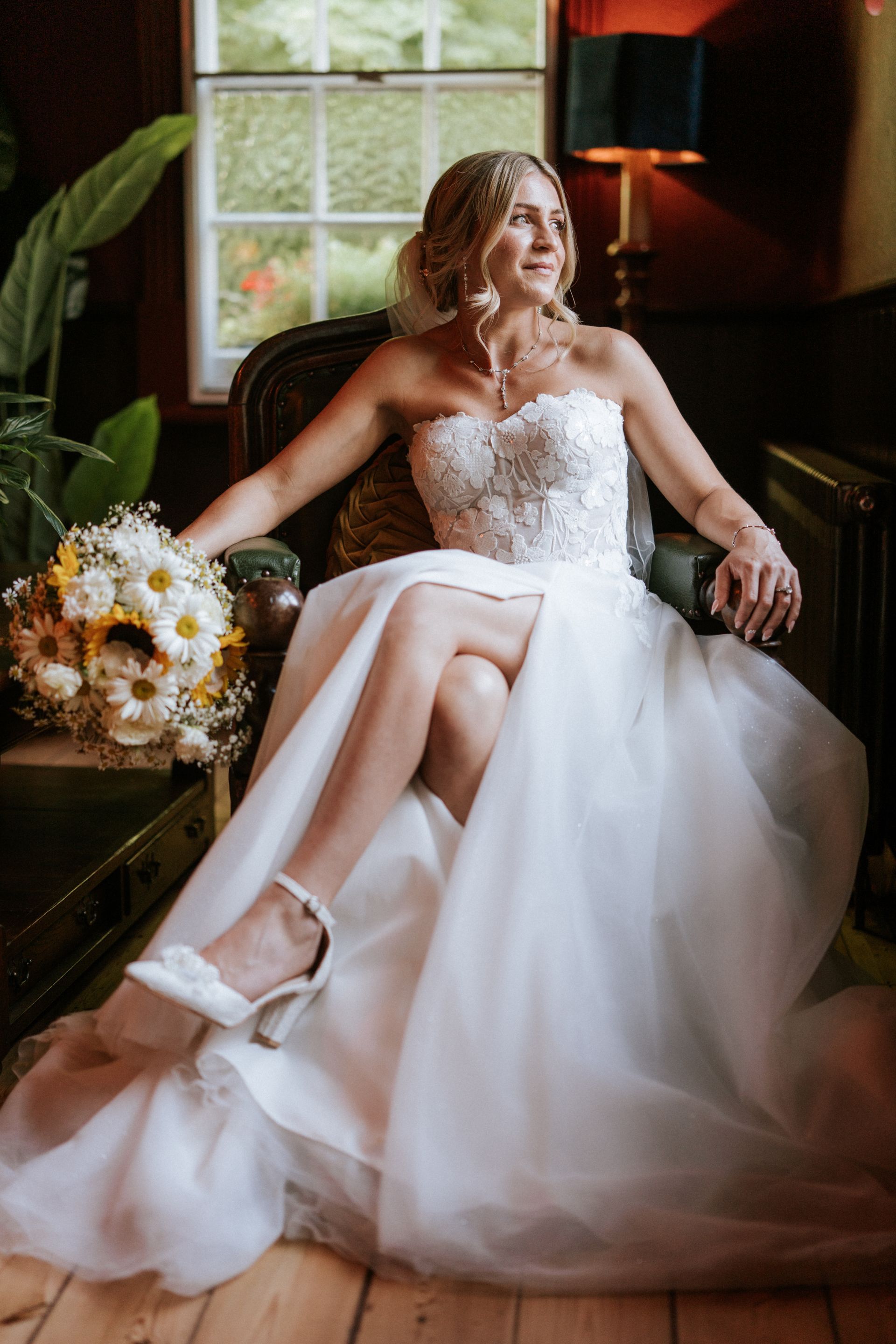 Bride in a white wedding gown seated in a dark room by a window, holding a bouquet and looking off-camera