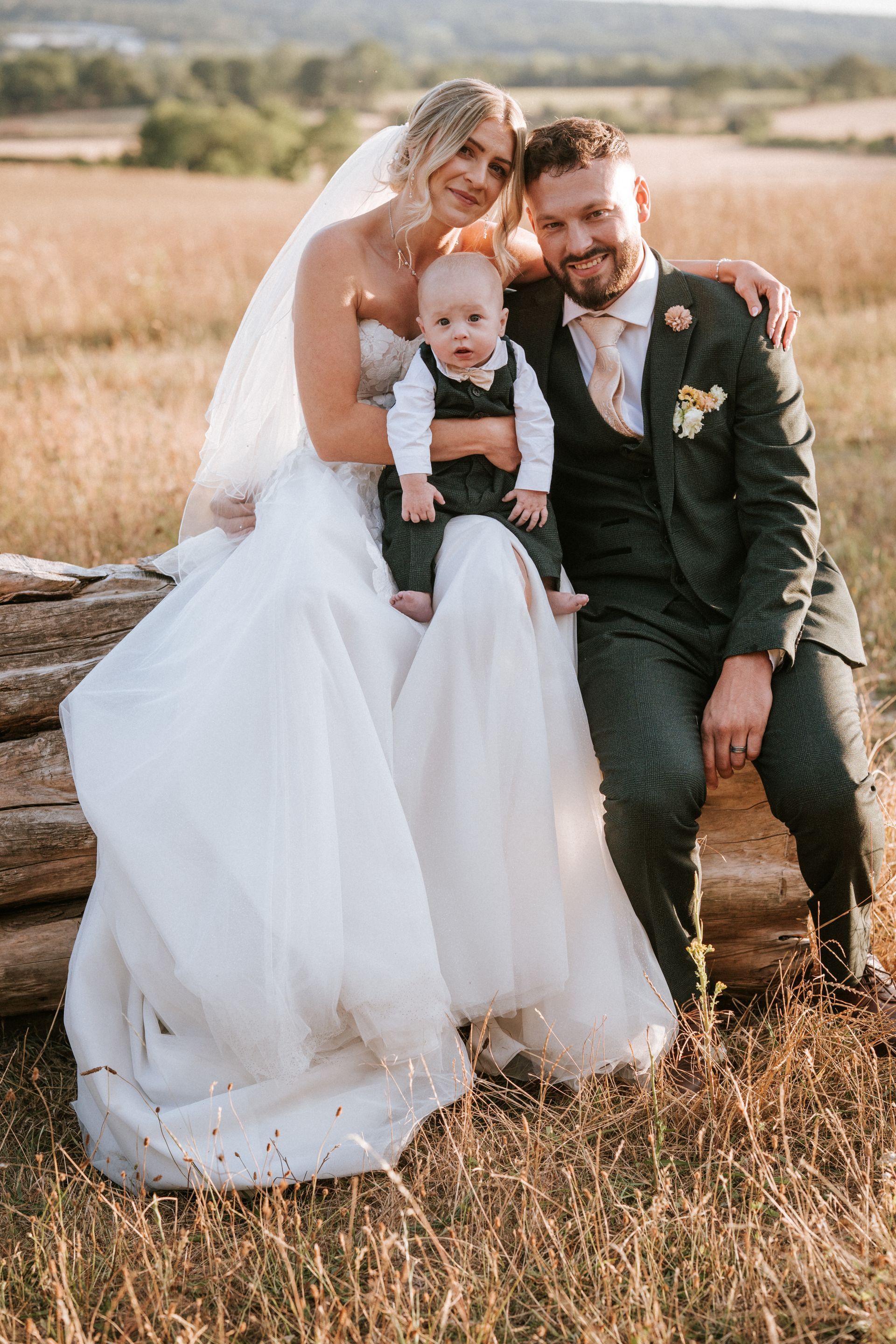 Bride, groom, and baby sitting together in a golden field, smiling at the camera.