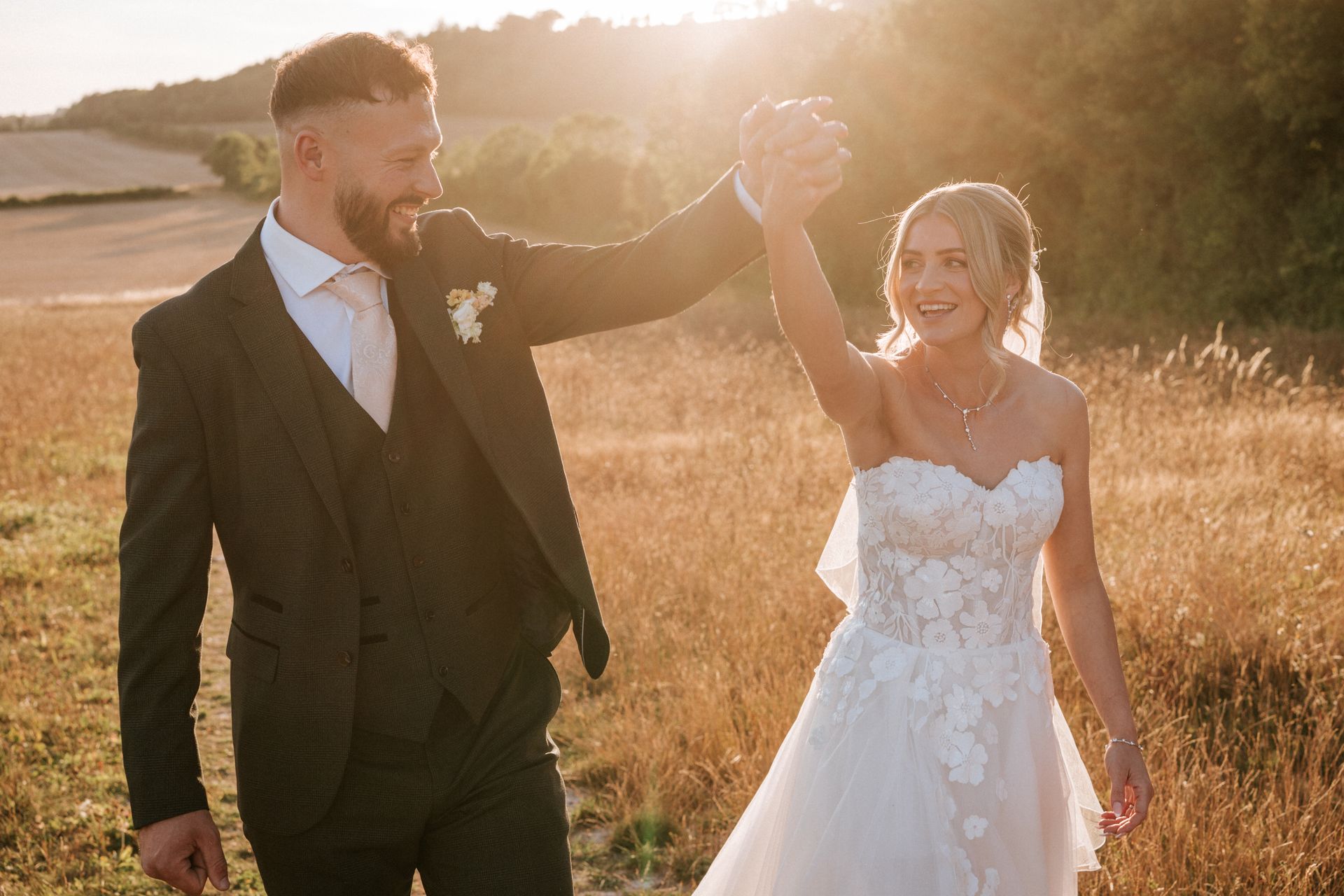 Bride and groom embracing on a sunlit woodland path, bride in white lace gown and veil holding a bouquet.