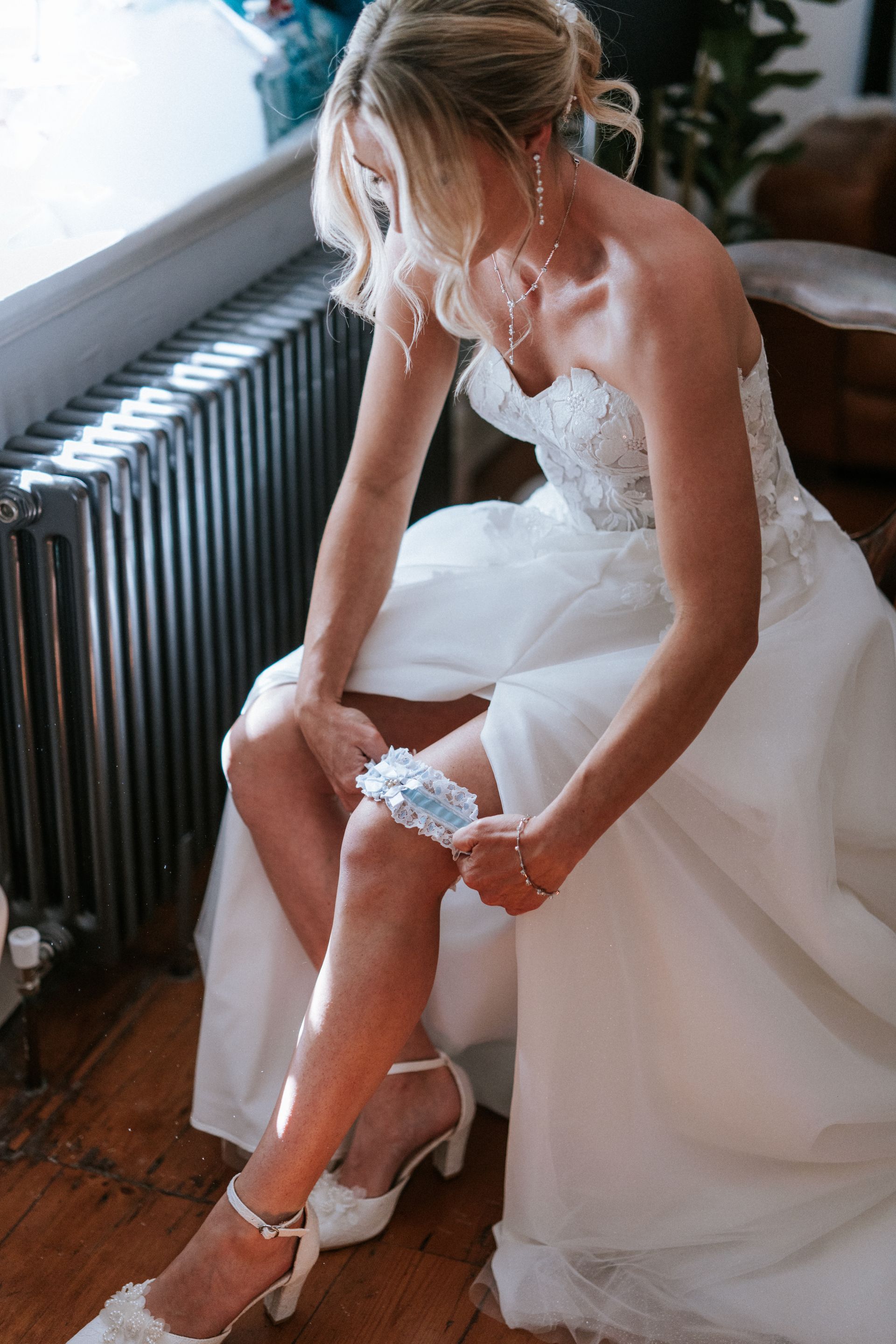 Bride adjusting her garter while sitting in a white wedding dress near a window and radiator